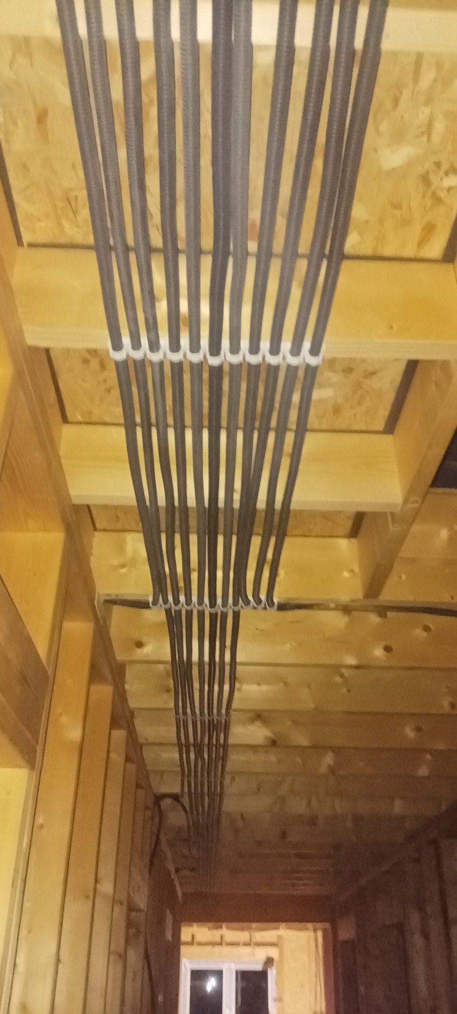 Równolegle ułożone, szare rury instalacyjne przymocowane białymi opaskami do drewnianej konstrukcji stropu. Widok z dołu na instalację w surowym wnętrzu.