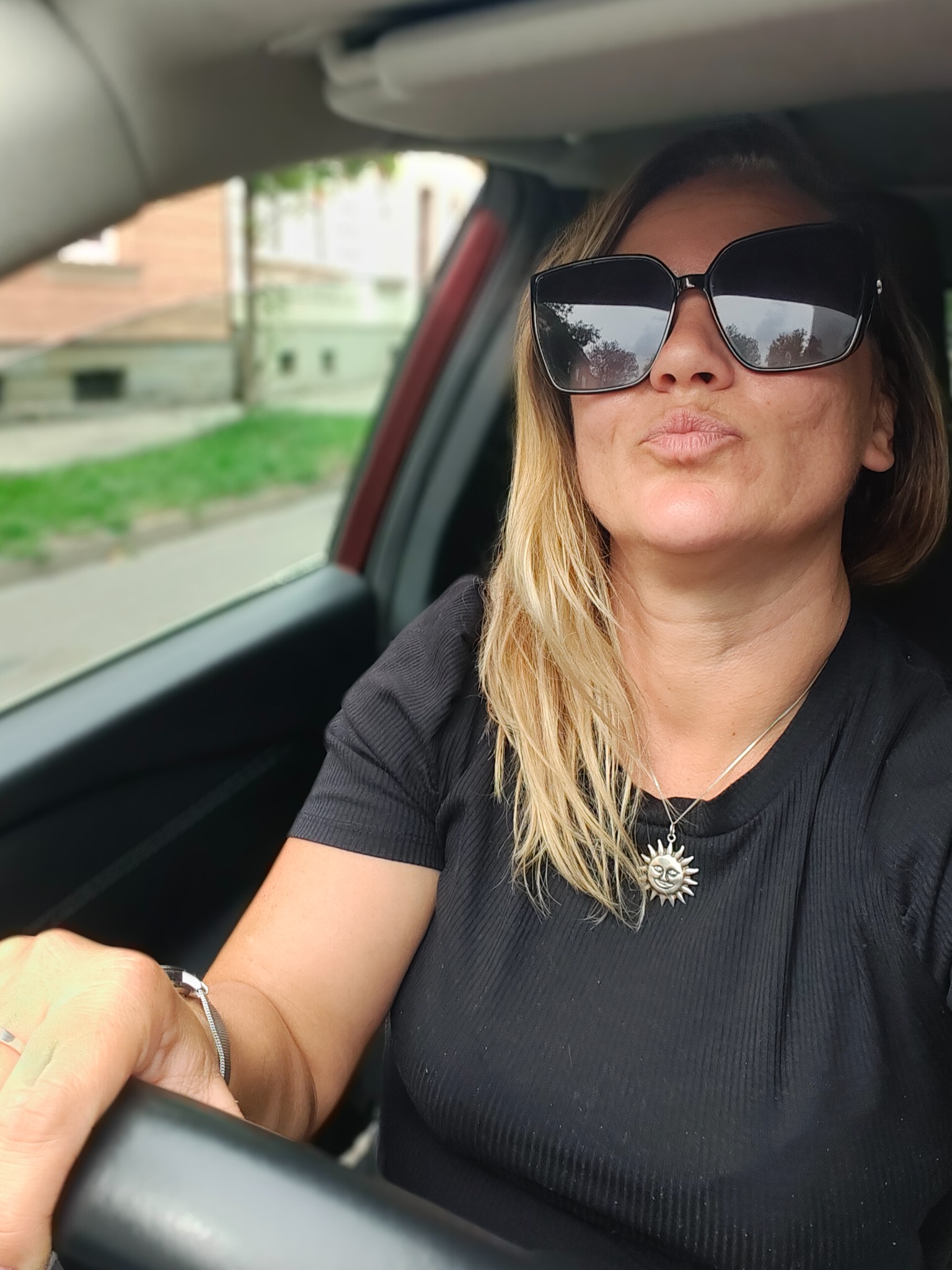 Uśmiechnięta blondynka w czarnych okularach siedzi za kierownicą czerwonego samochodu, robiąc selfie z dzióbkiem. W tle widoczny budynek i zieleń.