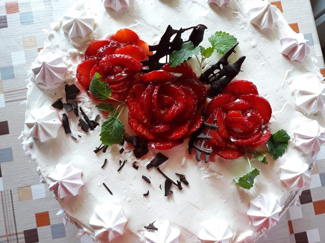 Tort z kremem, udekorowany truskawkowymi różami, czekoladą i bezami. Widok z góry, na tle wzorzystego obrusa.