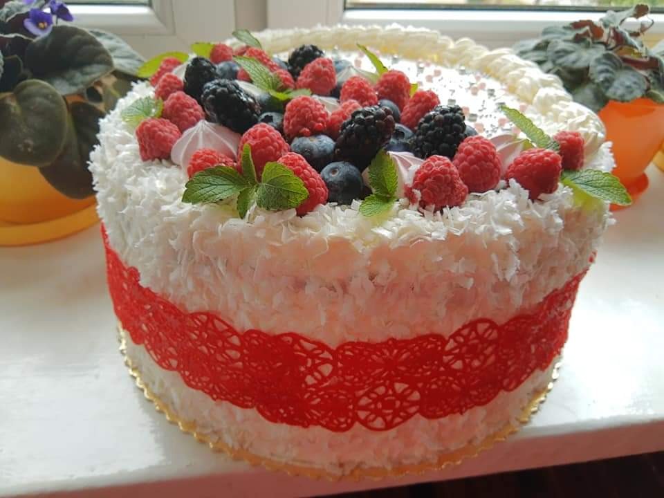 Tort z bitą śmietaną, kokosem, malinami, jeżynami i borówkami, udekorowany czerwoną koronką, na tle okna i kwiatów doniczkowych.