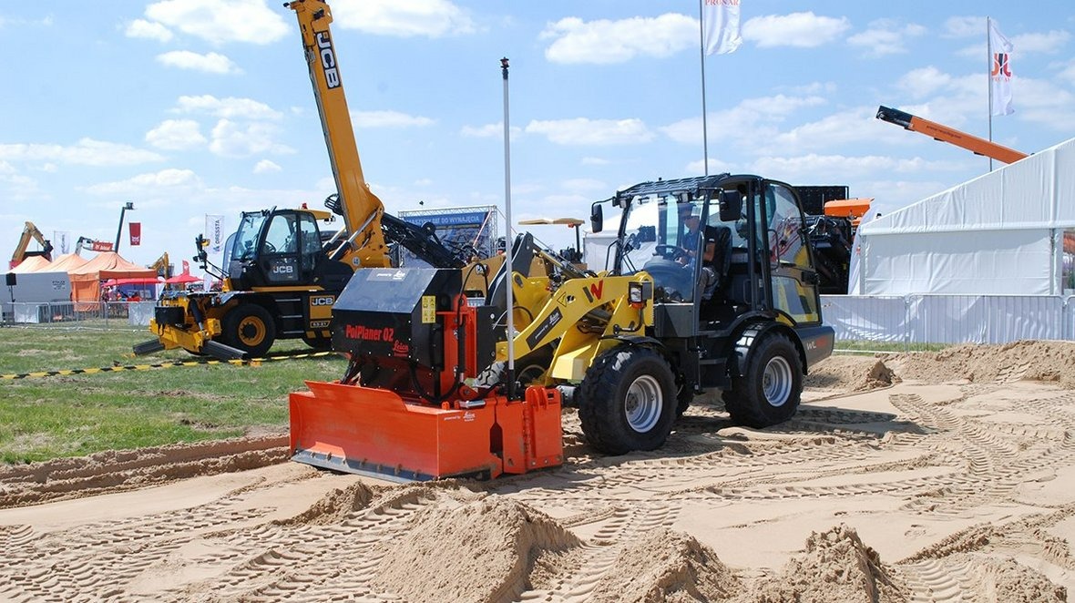 Żółta ładowarka z pomarańczowym osprzętem PolPlaner 02 na piaszczystym podłożu. W tle inne maszyny budowlane i namioty targowe pod błękitnym niebem.