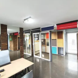 Ekspozycja różnorodnych rolet i żaluzji w salonie: rolety dzień noc, aluminiowe, drewniane. Widoczne próbki kolorów i faktur. Stonowana kolorystyka wnętrza.