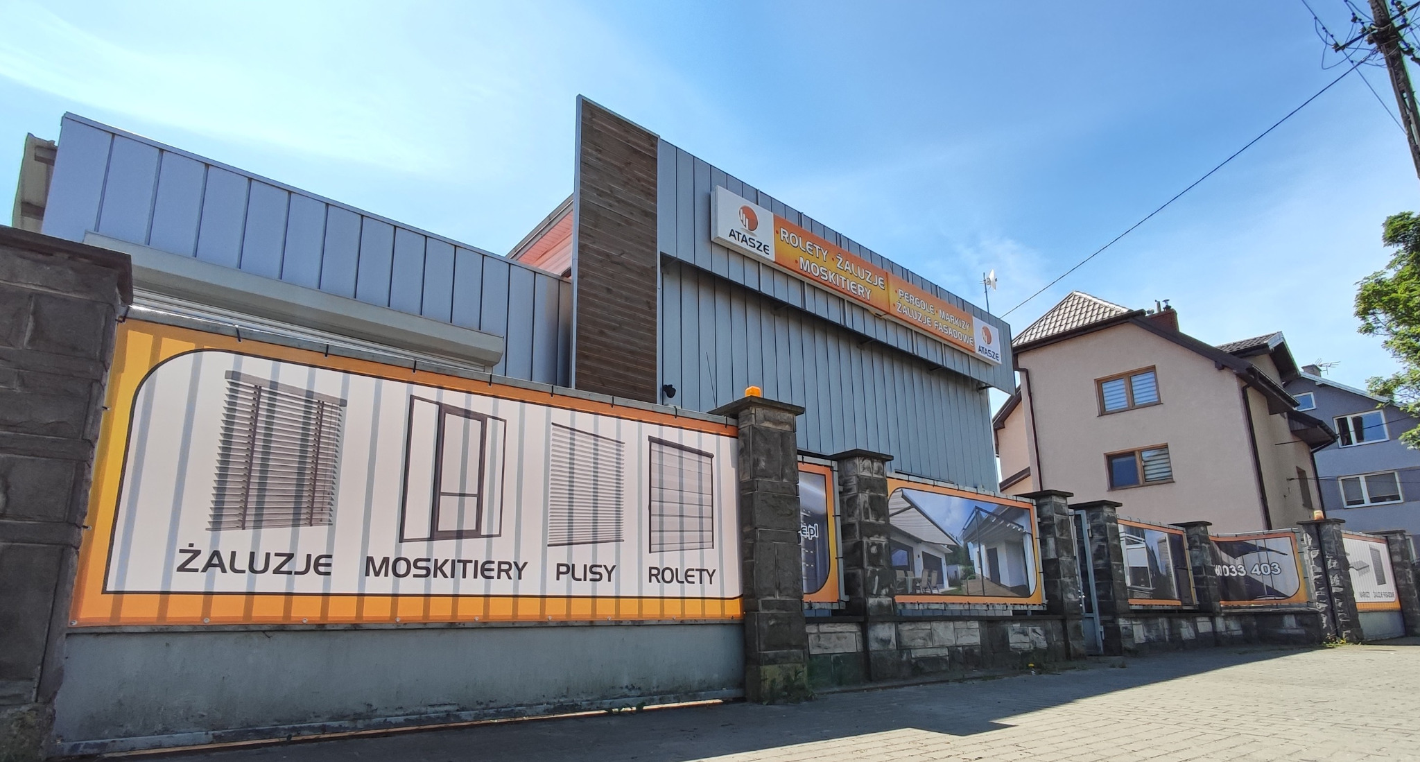 Reklama firmy Atasze na ogrodzeniu z żaluzjami, moskitierami, plisami i roletami. Budynek firmy z logo i nazwą na górze. Słoneczny dzień.