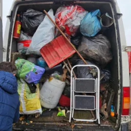 Załadowany van z workami, łopatą i drabiną. Widoczny fragment osoby w niebieskiej kurtce. Wywóz odpadów po pracach ogrodowych lub opróżnianiu.