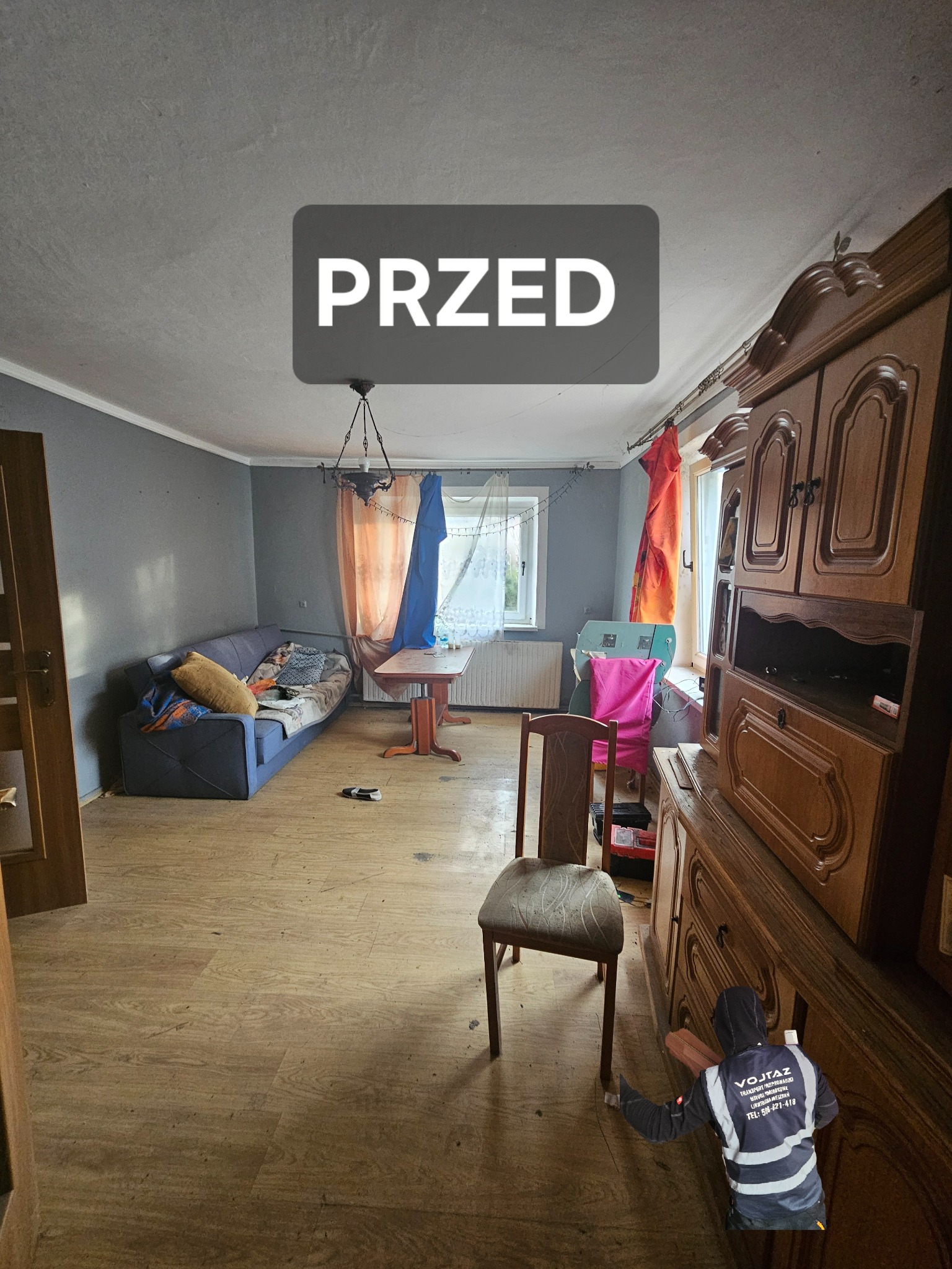 Pokój z drewnianą podłogą i meblami przed opróżnianiem. Widoczna sofa, stół, krzesło i duża szafa. Na zdjęciu napis 'PRZED'.