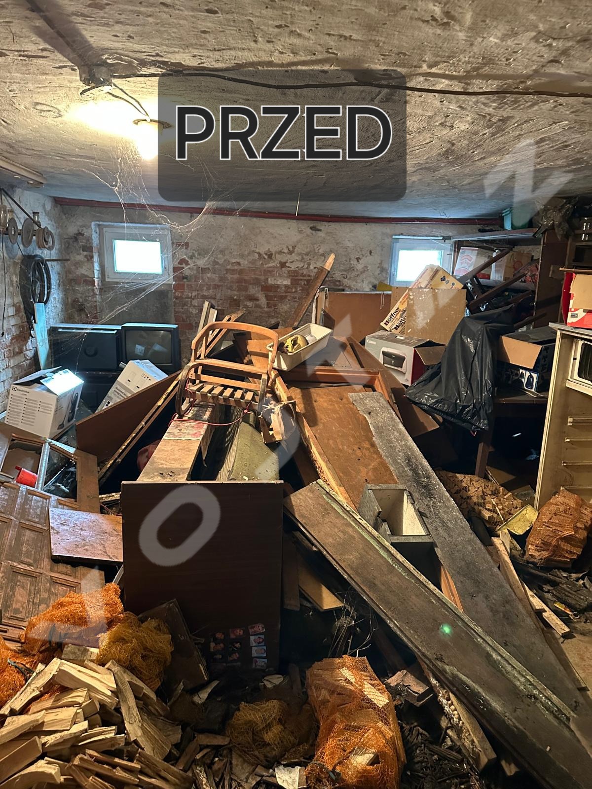 Zagracony stary pokój z meblami, sprzętami i odpadami. Na zdjęciu napis 'PRZED'. Widoczne okna, pajęczyny i stara ceglana ściana.