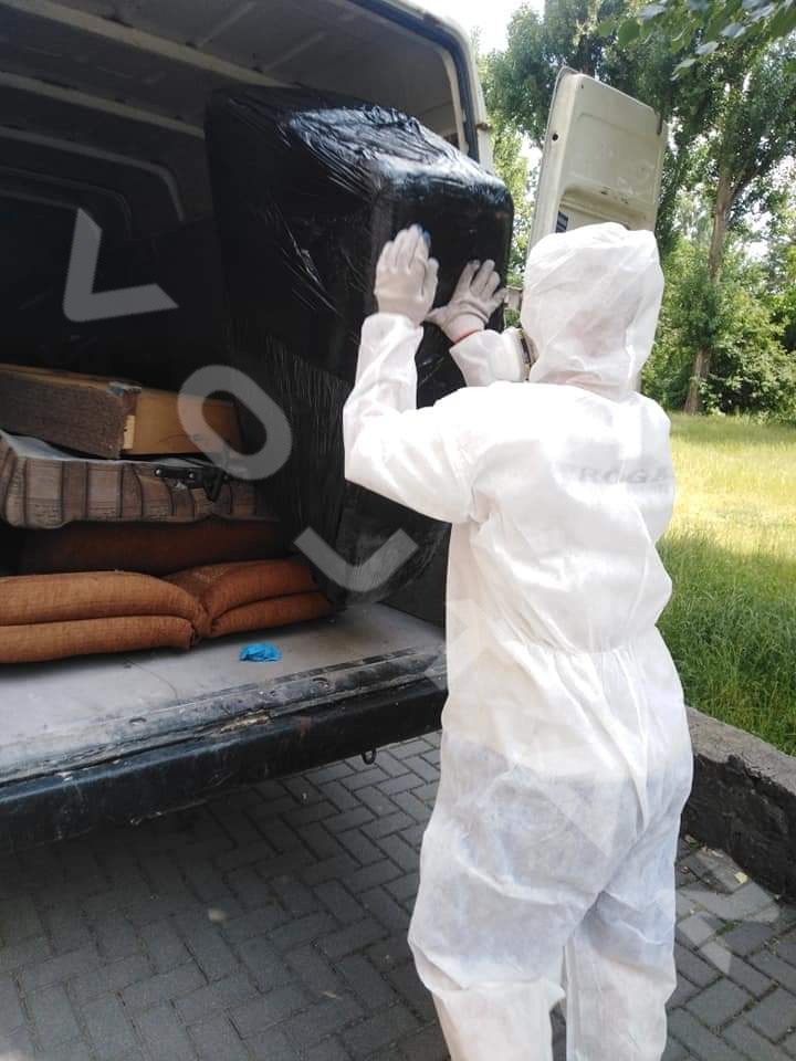 Pracownik w kombinezonie ochronnym wkłada zapakowany mebel do furgonetki. W tle widoczne inne meble i drzewa. Usługa opróżniania.