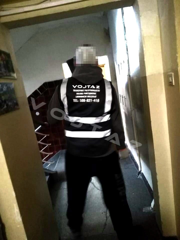 Pracownik firmy VOJTAZ w odblaskowej kamizelce w trakcie opróżniania mieszkania na klatce schodowej. Widoczne ściany i schody w tle.