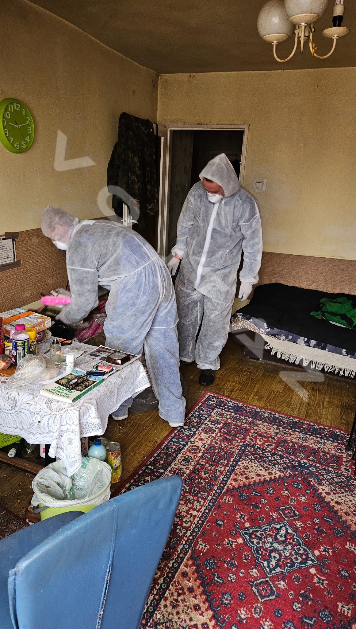 Dwóch mężczyzn w kombinezonach ochronnych porządkuje zagracone mieszkanie. Na stole leżą różne przedmioty, w tle widoczny dywan i meble. Praca w toku.