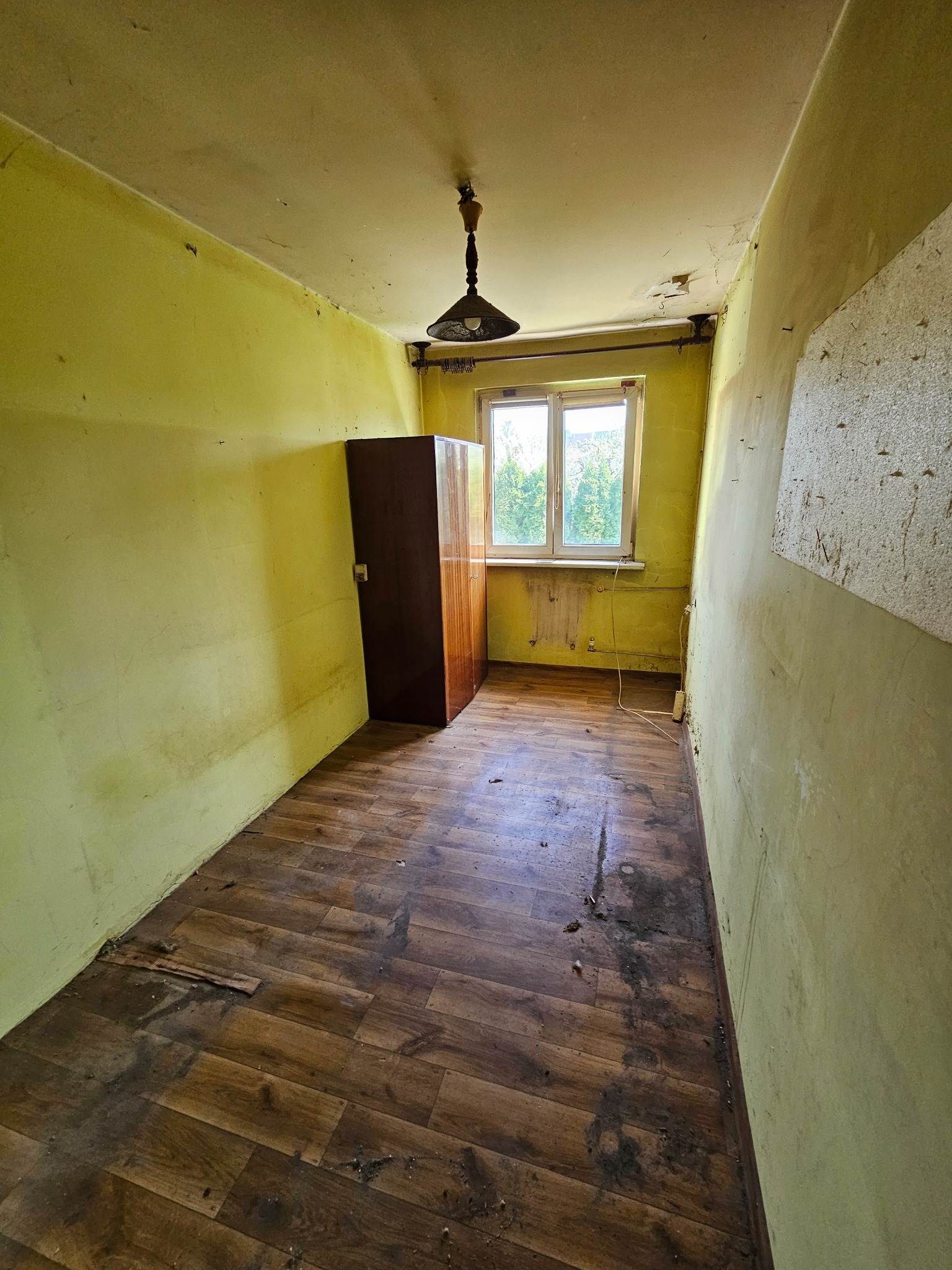 Pokój przed opróżnianiem w Prudniku: żółte ściany, brudna podłoga, szafa i okno. Widoczne ślady użytkowania i zaniedbania. Perspektywa wnętrza z lampą.