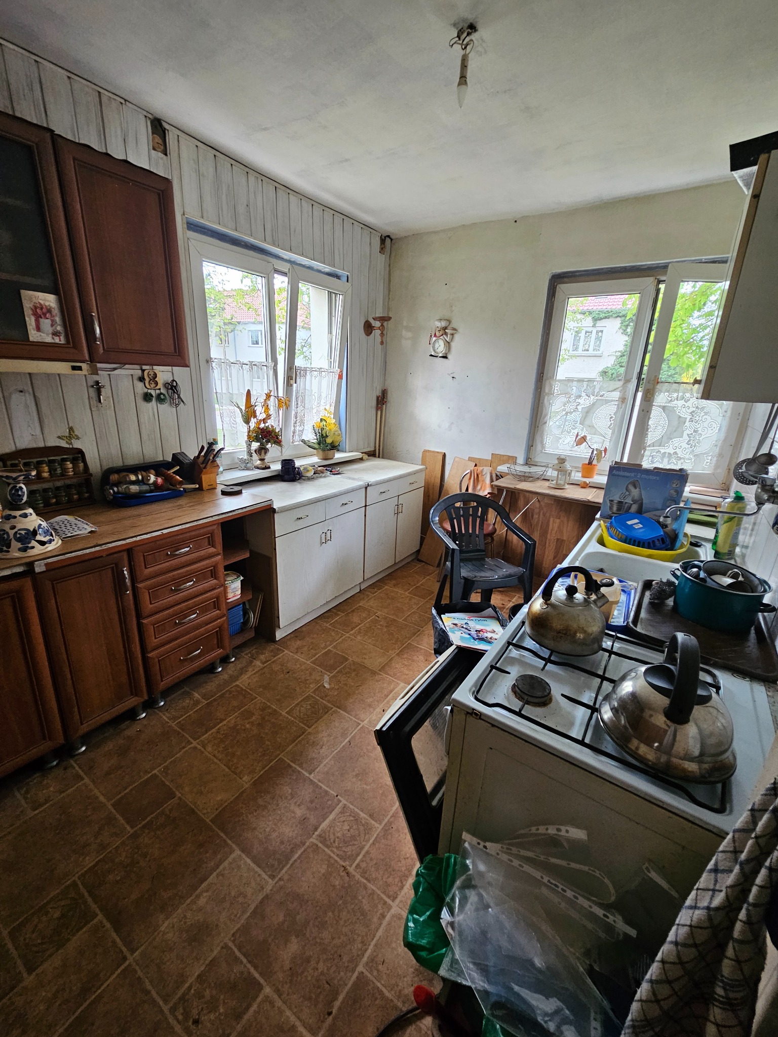 Kadr z wnętrza kuchni przed remontem: stare meble, kuchenka z czajnikami, okna z firankami, brązowa podłoga. Widok na zaniedbane pomieszczenie.