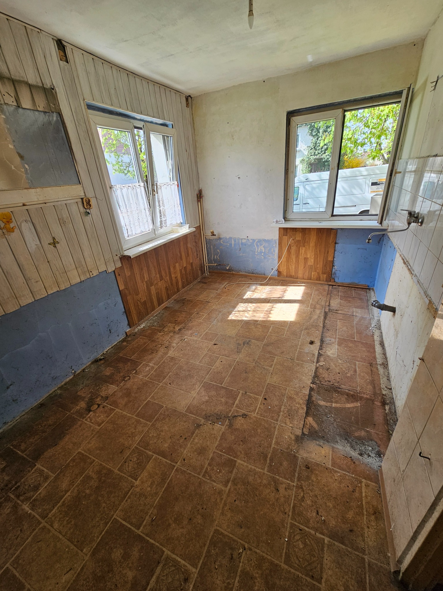 Kadr z wnętrza mieszkania w trakcie opróżniania: zdemontowane panele ścienne, stara podłoga z płytek, widoczne ślady po instalacjach. W tle okno z widokiem na samochód dostawczy.