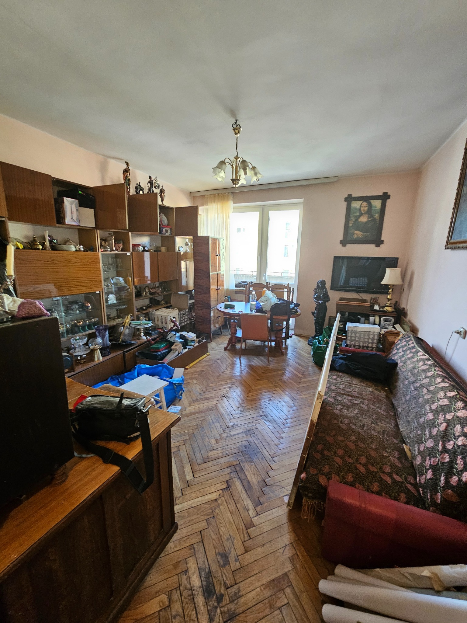 Zastawiony salon z parkietem w jodełkę, meblościanką pełną bibelotów i sofą. Widoczne przedmioty przygotowane do pakowania, sugerujące etap opróżniania mieszkania.