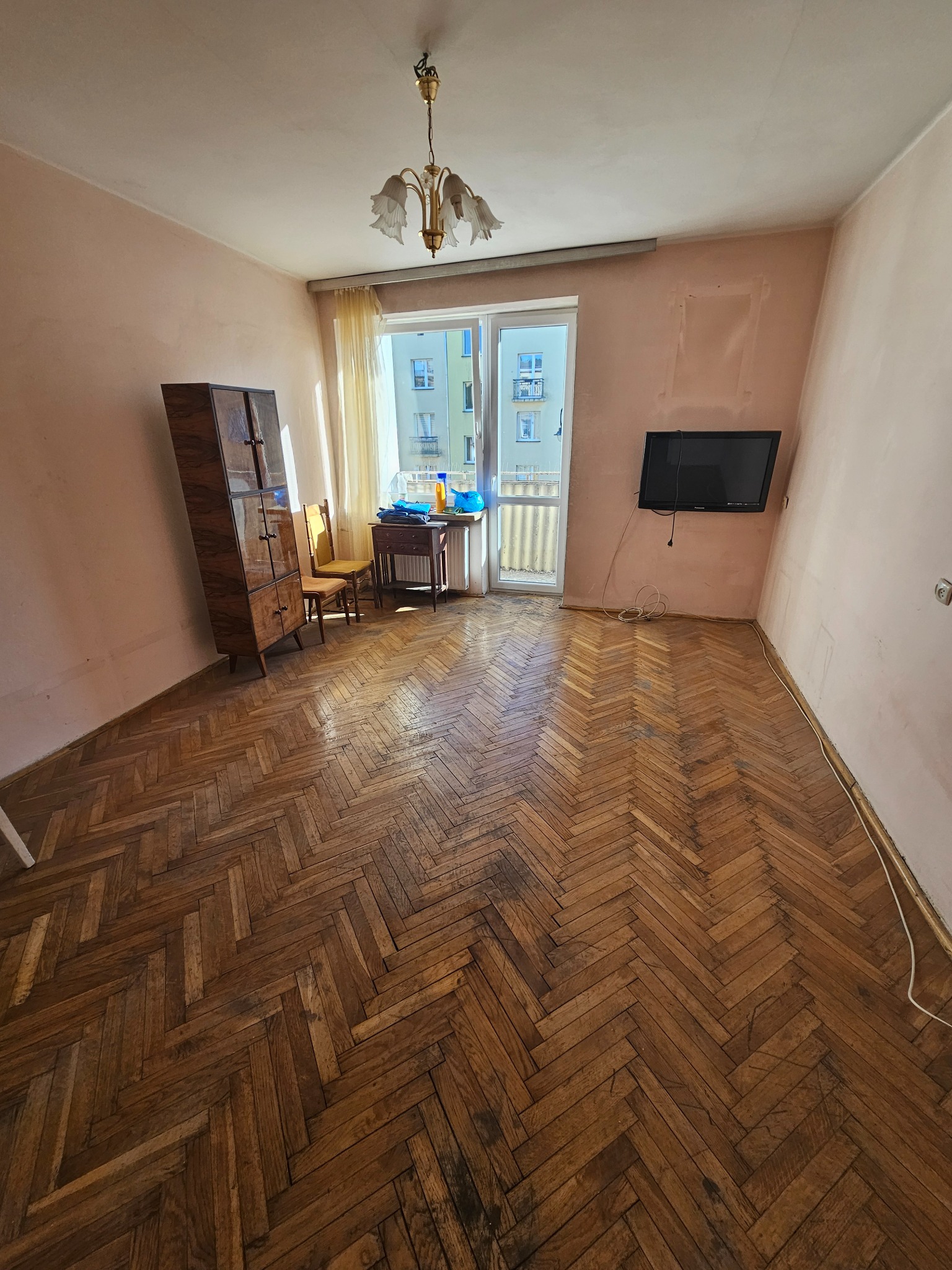 Pusty pokój z parkietem w jodełkę, szafą, krzesłami i telewizorem na ścianie. Widoczne ślady po remoncie, okno balkonowe z widokiem na budynek.