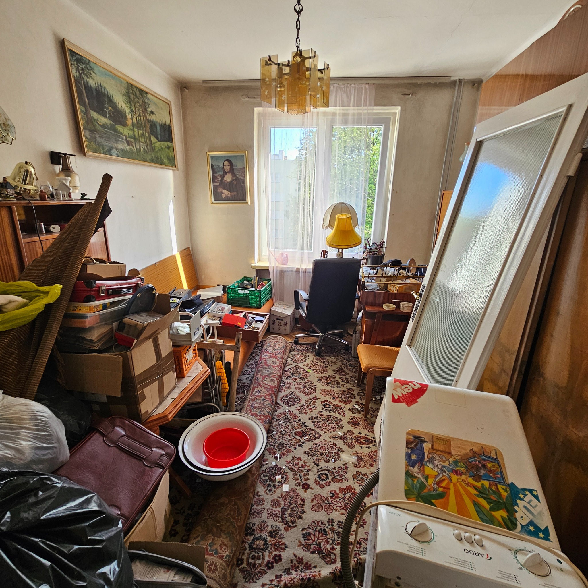 Zagracony pokój z meblami, kartonami i sprzętami AGD. Na ścianie wiszą obrazy, a przy oknie stoi biurko z krzesłem. Widoczna stara pralka z naklejkami.