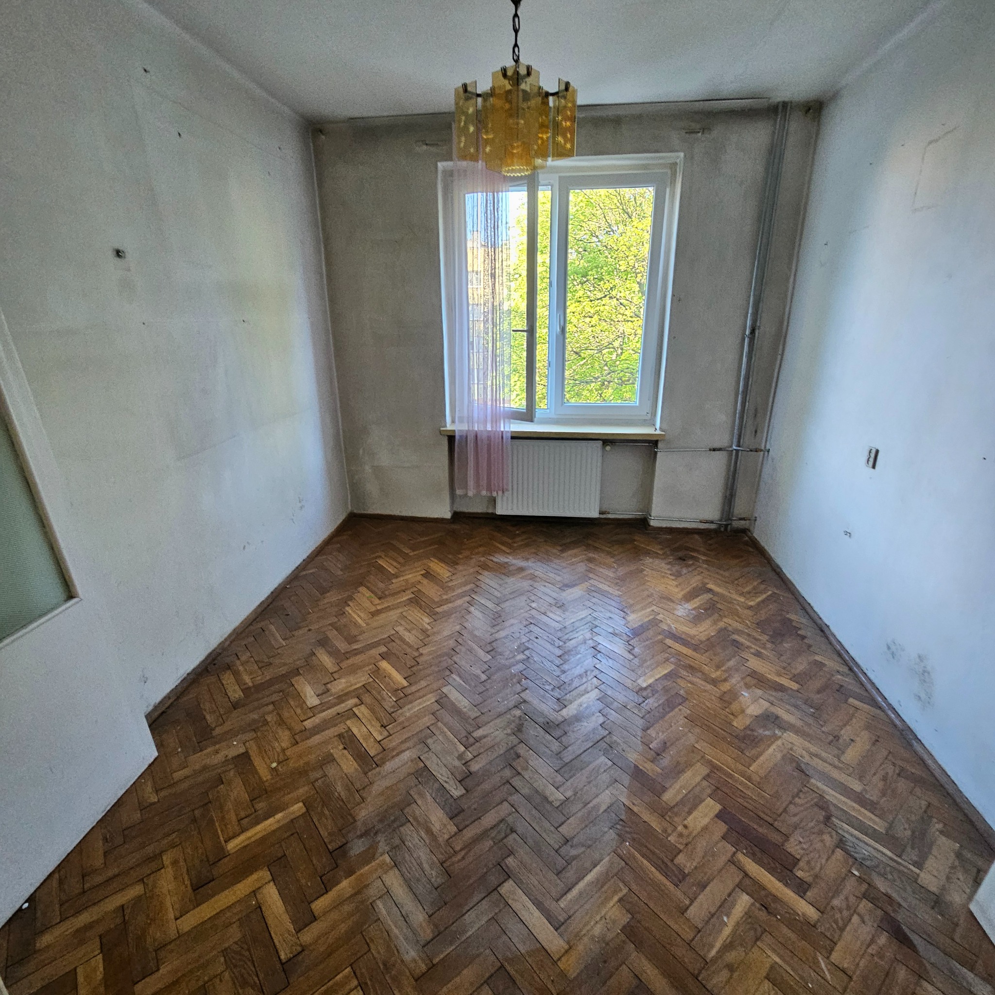 Puste, zaniedbane wnętrze z parkietem w jodełkę, oświetlone przez okno z widokiem na zieleń. Stara lampa wisząca dodaje charakteru. Ściany wymagają odświeżenia.