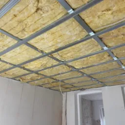 Sufit podwieszany z metalową konstrukcją wypełnioną żółtą wełną mineralną, widok z dołu, surowe ściany w tle, wnętrze w trakcie remontu.