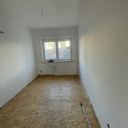 Pokój po gładziowaniu ścian, z oknem i podłogą z płyt OSB. Na podłodze leży kij teleskopowy. Widoczne gniazdka elektryczne.