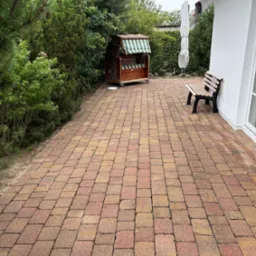 Ułożona kostka brukowa w odcieniach czerwieni i brązu na posesji. W tle widoczna drewniana ławeczka przy białej ścianie domu z strzechą i kosz plażowy.