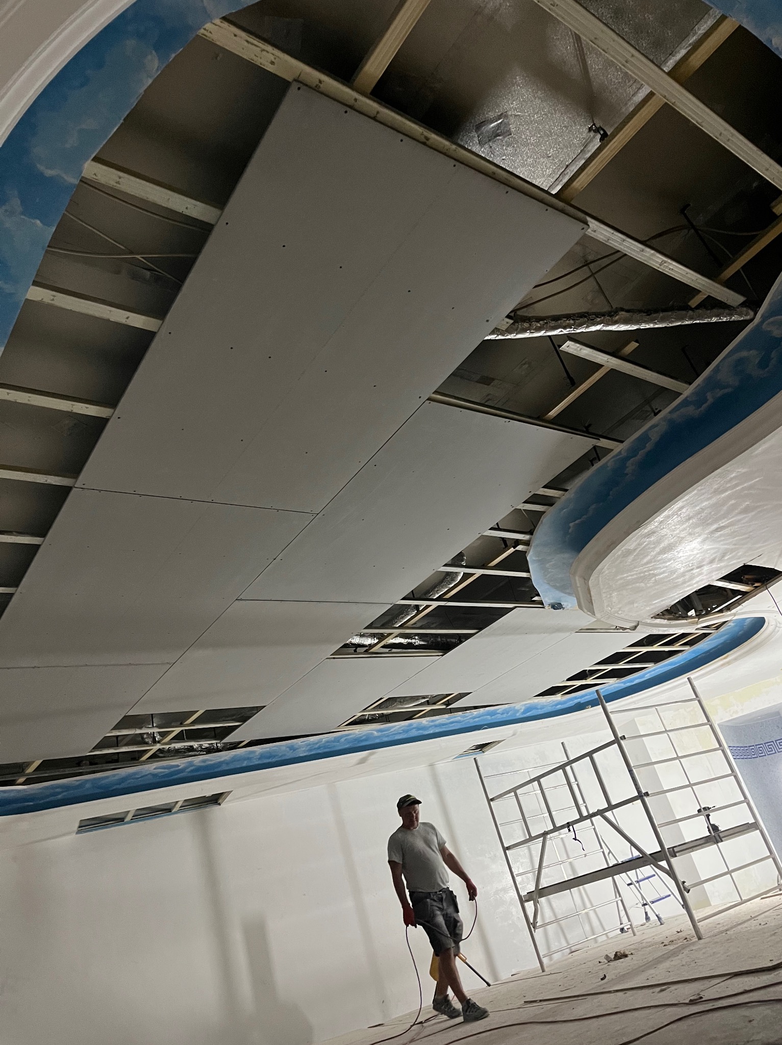 Montaż płyt karton-gips na suficie podwieszanym o nietypowym kształcie. Widoczny pracownik oraz rusztowanie. Elementy dekoracyjne sufitu w kolorze niebieskim.