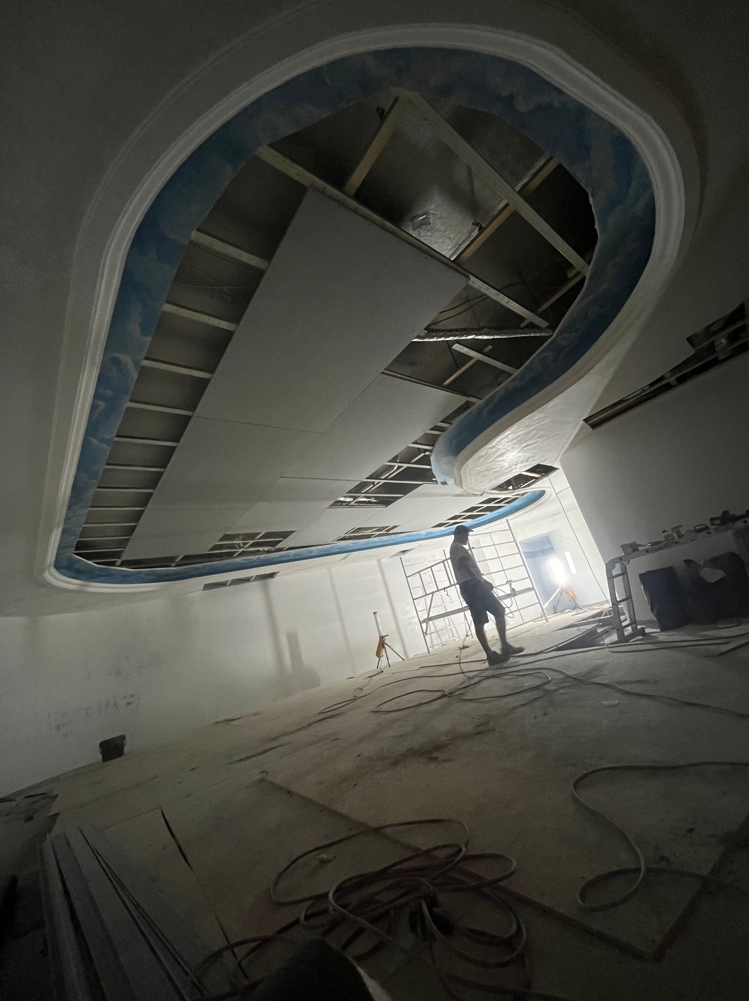 Sufit z płyty gipsowej z dekoracyjnym, niebieskim obramowaniem w trakcie montażu. Widoczny pracownik na rusztowaniu i elementy konstrukcji. Ujęcie z dołu.