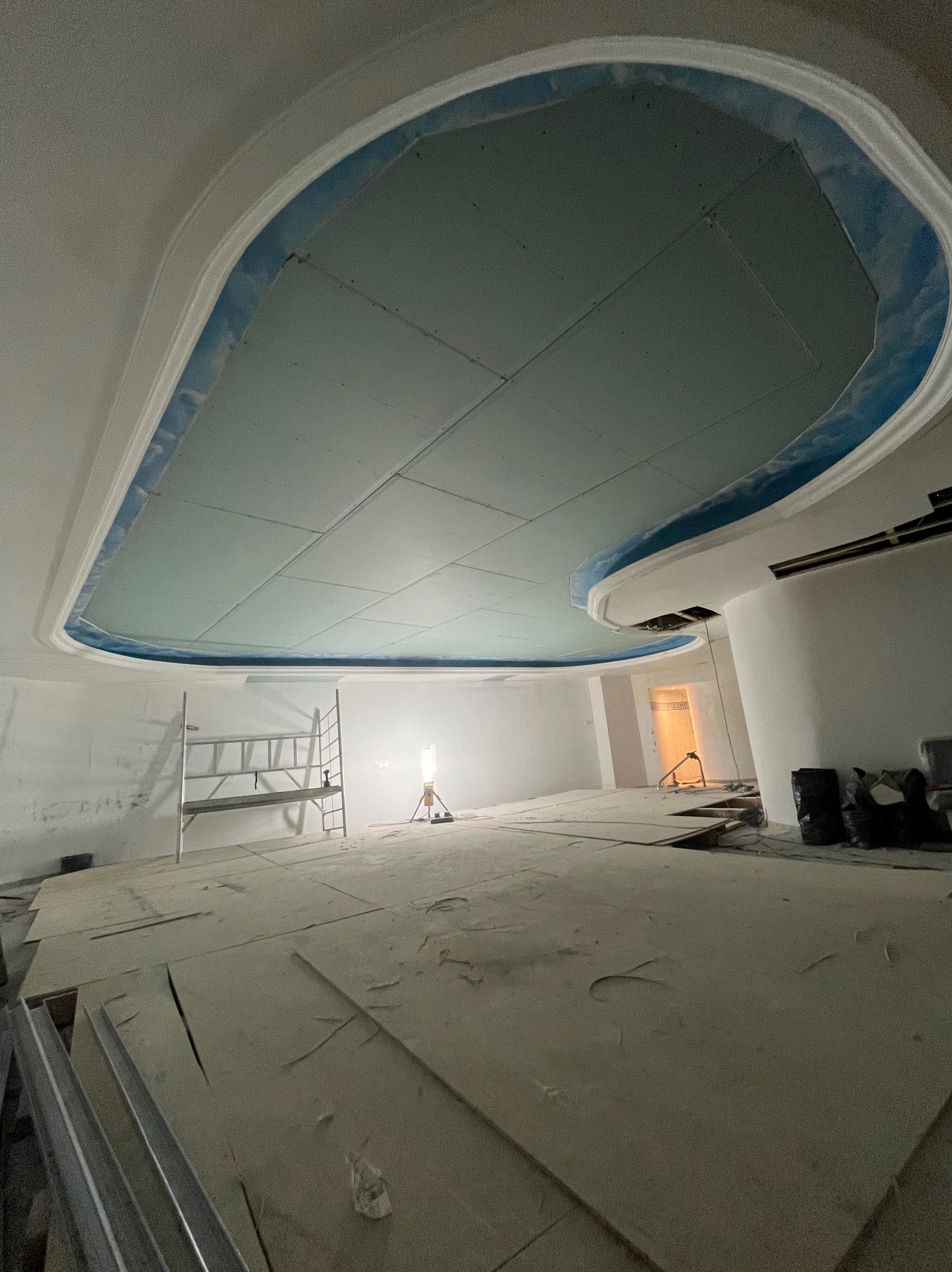 Sufit podwieszany z płyt karton-gips w trakcie montażu, widoczne profile, szpachlowanie łączeń, oświetlenie budowlane. W tle drabina i elementy wykończeniowe.