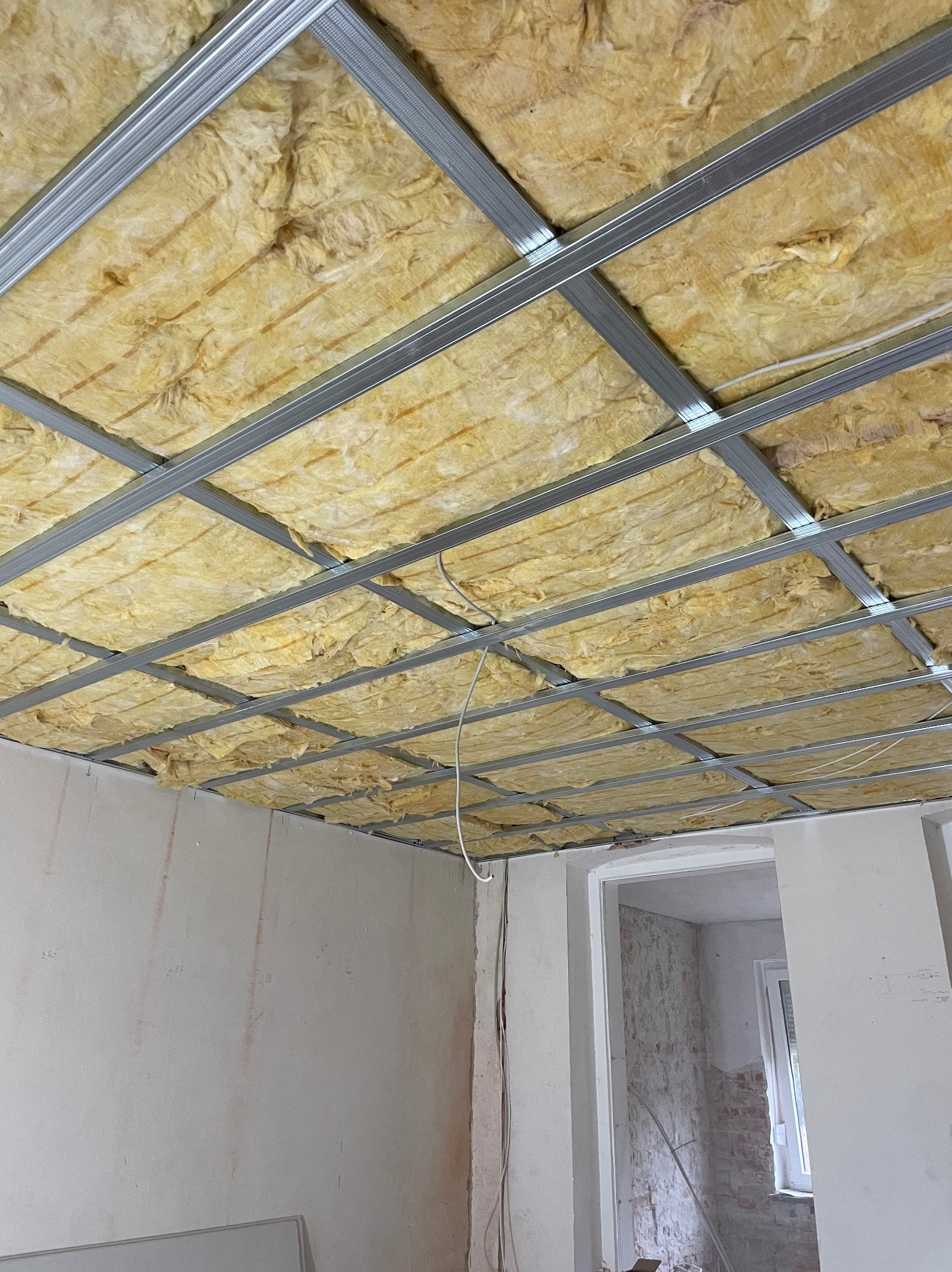 Sufit podwieszany z metalową konstrukcją wypełnioną żółtą wełną mineralną, widok z dołu, surowe ściany w tle, wnętrze w trakcie remontu.