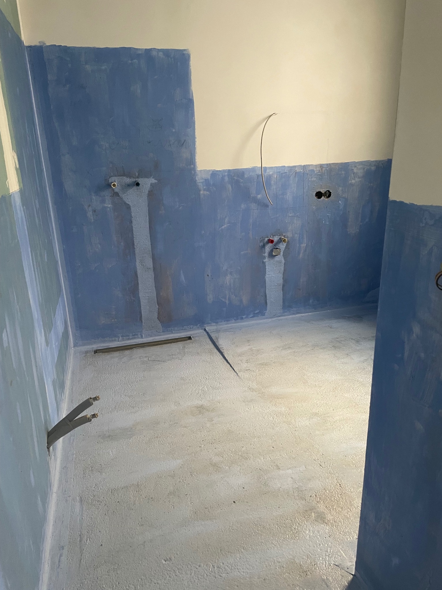 Przygotowana do wykończenia łazienka: ściany z niebieską hydroizolacją, widoczne rury i odpływ liniowy w podłodze. Surowy stan po pracach przygotowawczych.