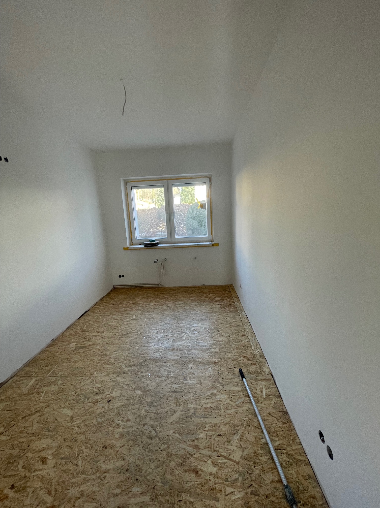 Pokój po gładziowaniu ścian, z oknem i podłogą z płyt OSB. Na podłodze leży kij teleskopowy. Widoczne gniazdka elektryczne.