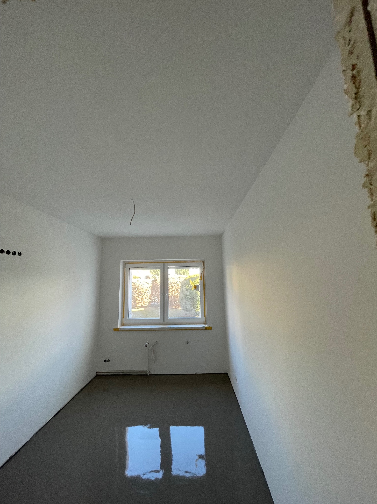 Wnętrze pokoju po wylaniu samopoziomującej wylewki; gładkie, szare podłoże odbija światło z okna. Ściany białe, przygotowane do dalszych prac wykończeniowych.