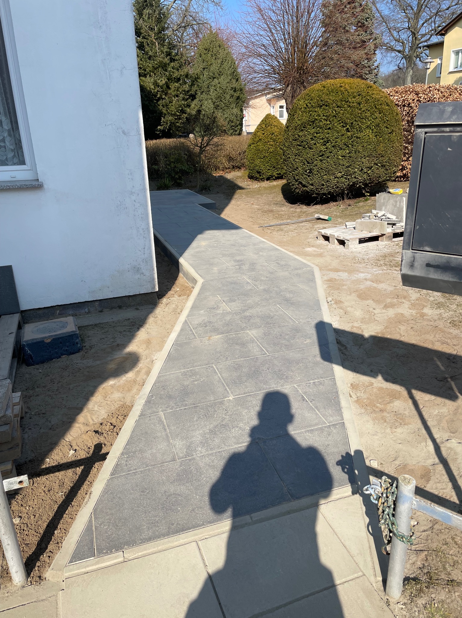 Ścieżka z szarych płyt ułożona w ogrodzie, obok biały budynek i zielone krzewy. Widoczny cień osoby wykonującej zdjęcie. Prace brukarskie w słoneczny dzień.