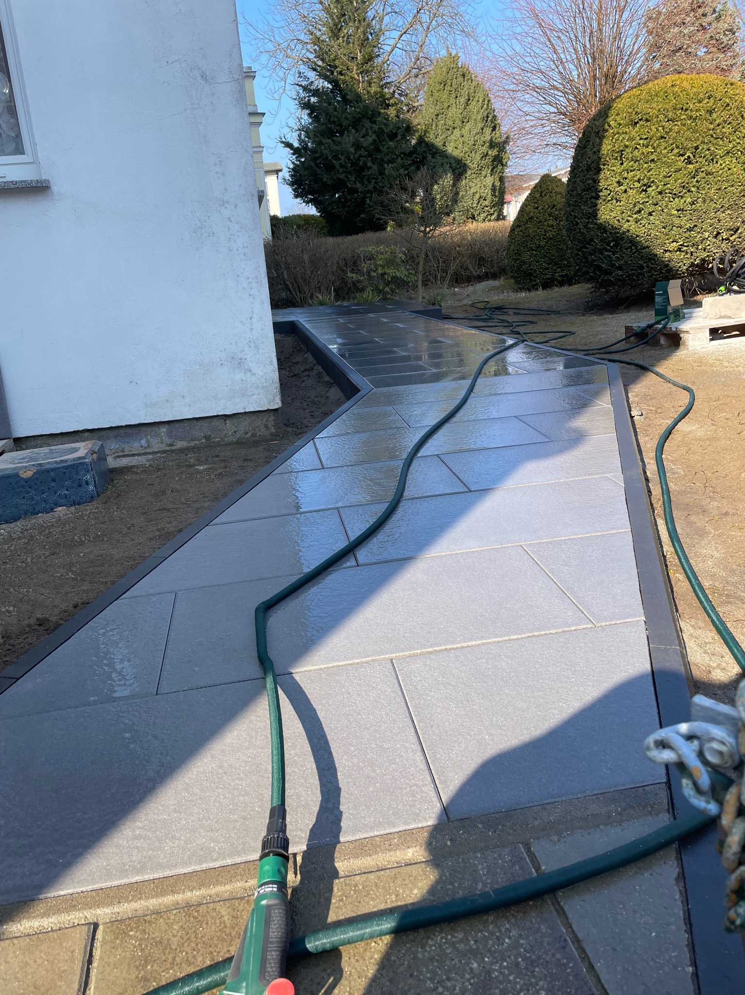 Świeżo ułożony chodnik z szarych płyt, obramowany czarnym obrzeżem, mokry od wody z węża ogrodowego. Widoczne narzędzia i fragment budynku w tle.