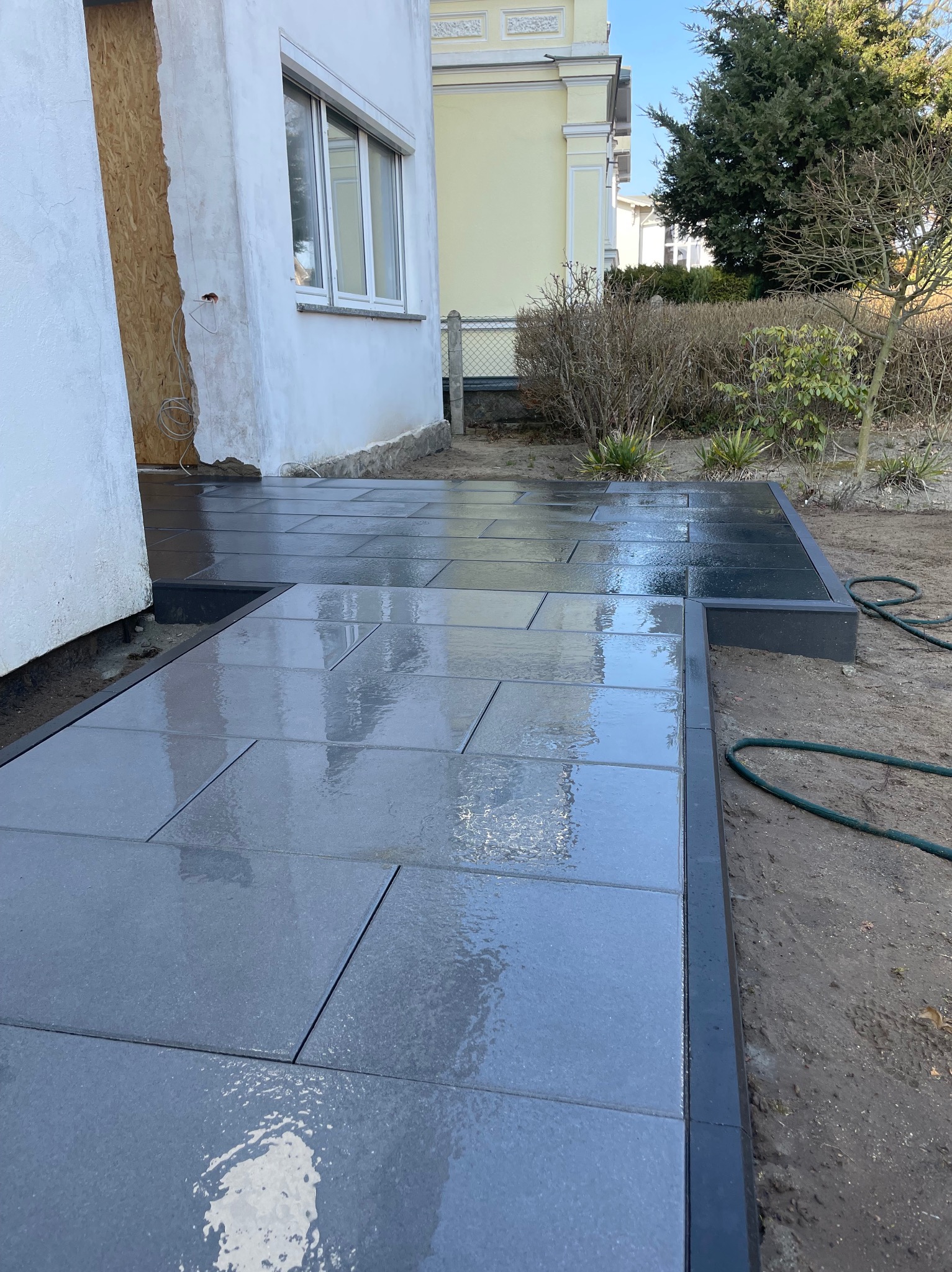 Nowa, ciemnoszara nawierzchnia z płyt kamiennych, ułożona wokół budynku z widocznymi śladami remontu. Błyszcząca powierzchnia, świeżo po deszczu.