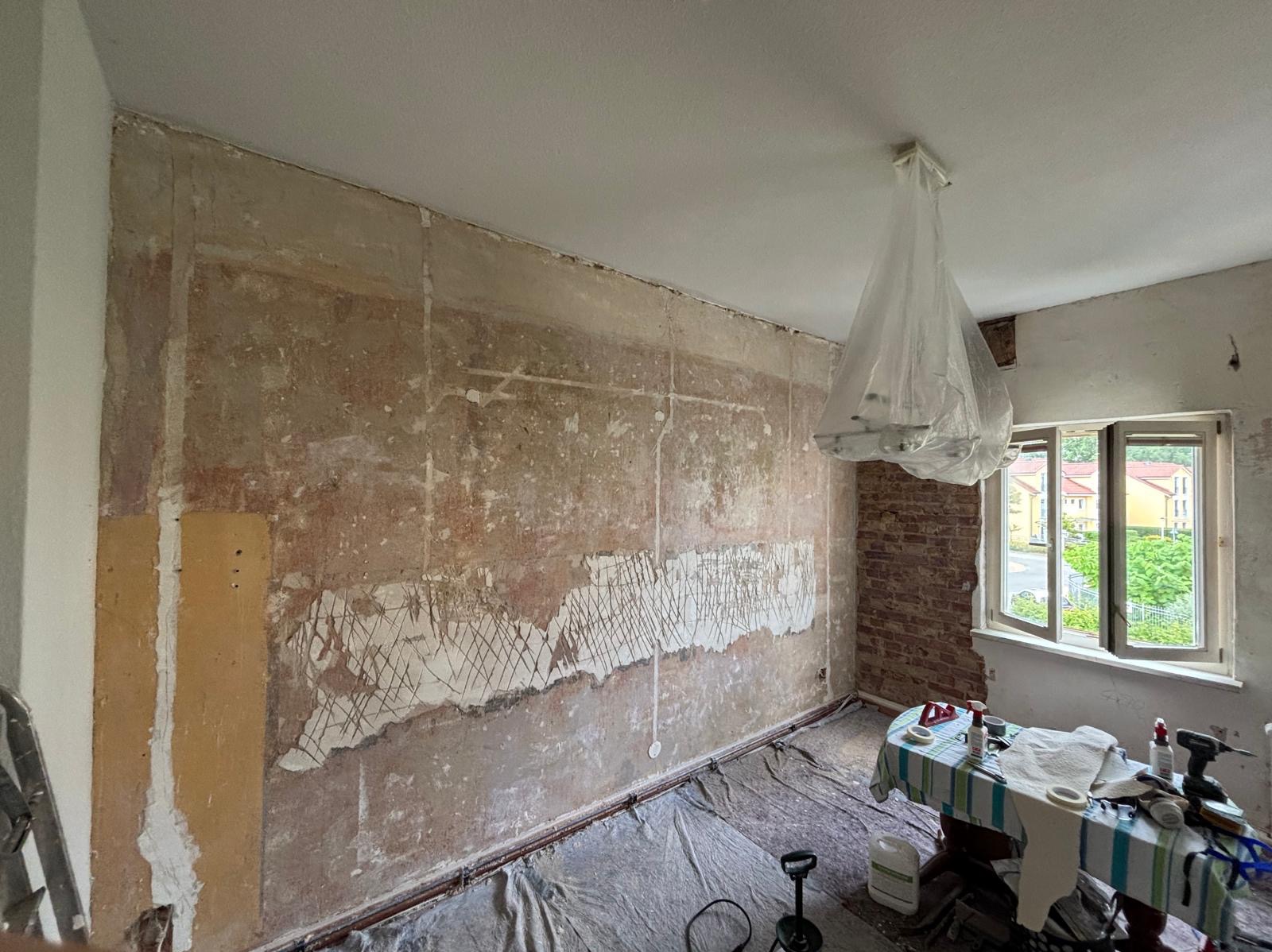Pokój w trakcie przygotowania do gładzi: zdarte stare powłoki, zarysowana siatka pod tynk, zabezpieczona lampa. Widoczne narzędzia i materiały na stole pod oknem.