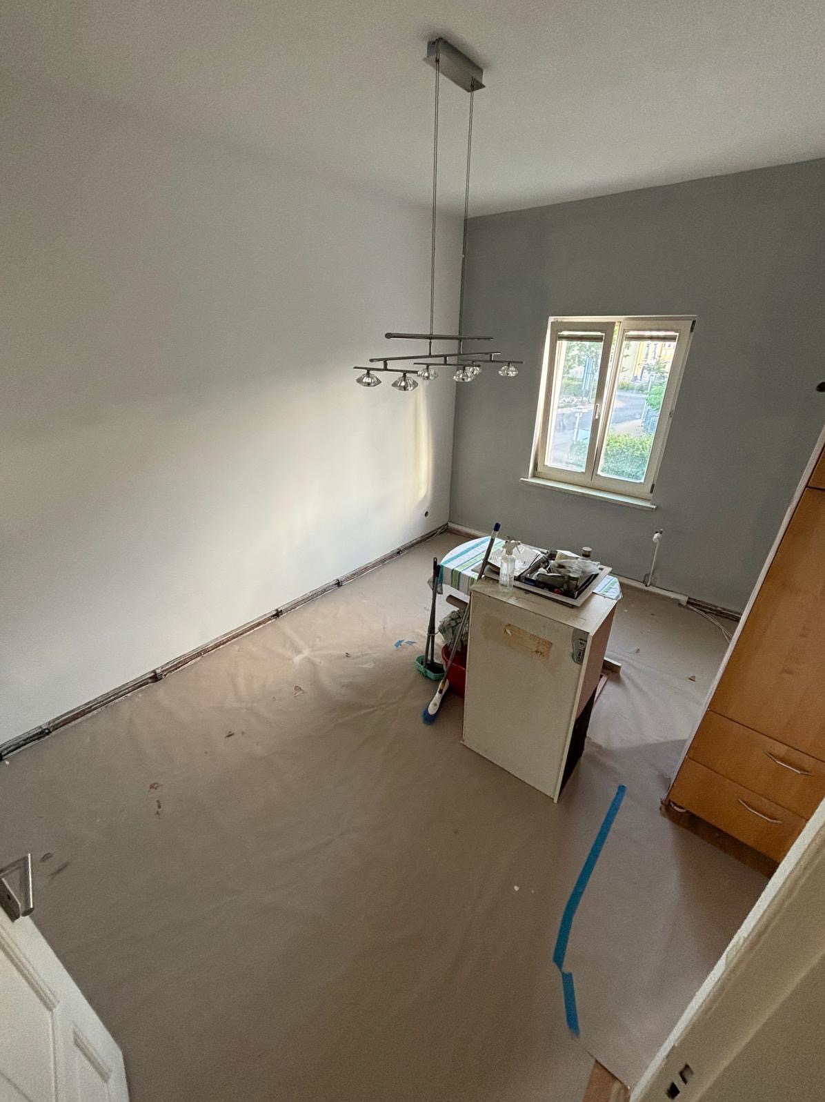 Pokój po malowaniu ścian na szaro, zabezpieczony folią malarską. Widoczny sprzęt malarski i lampa sufitowa. Ściany gładkie, jasne, z oknem w tle.