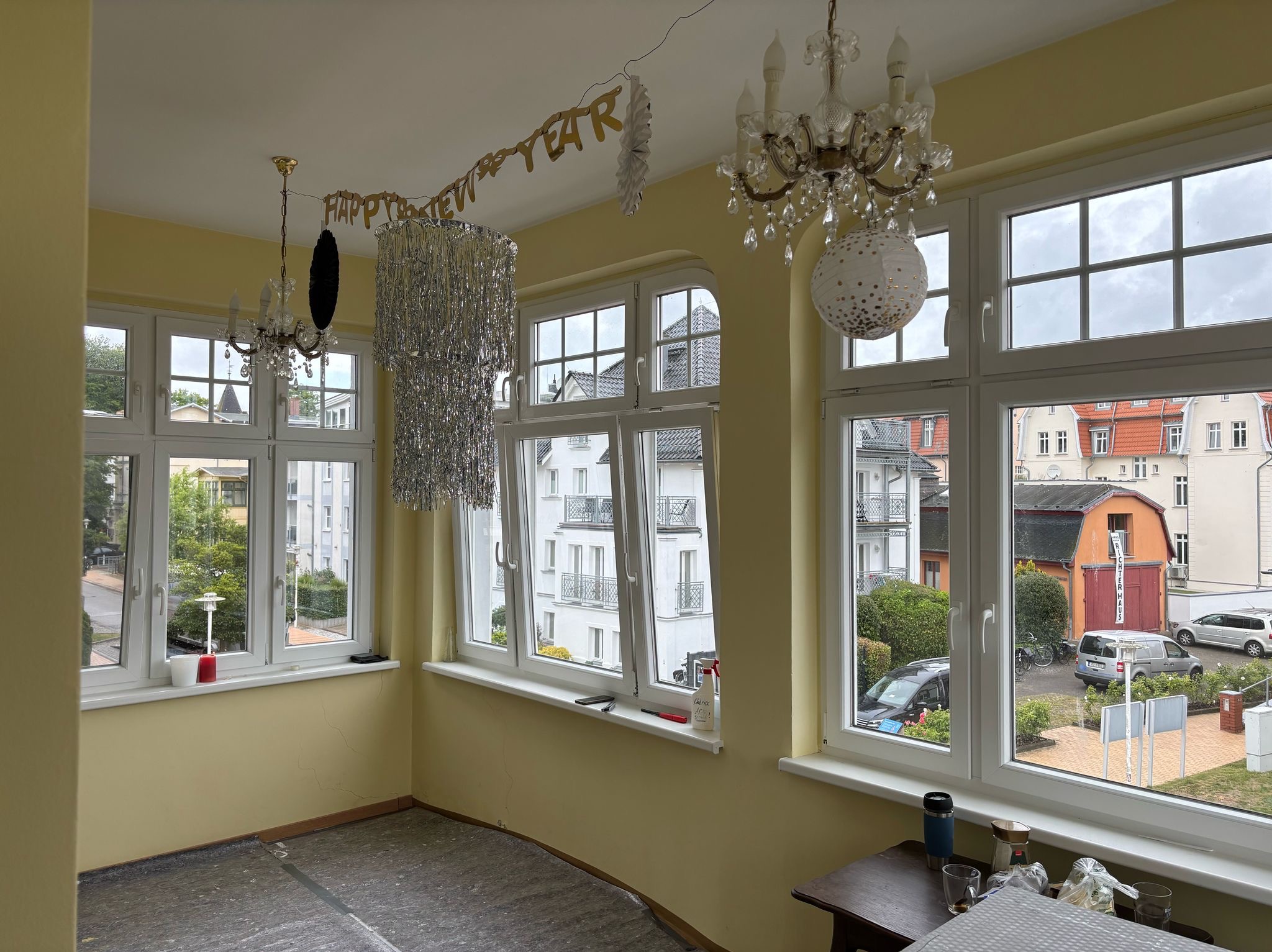 Żółte wnętrze pokoju z oknami, dekoracjami noworocznymi i widokiem na budynki. Podłoga zabezpieczona folią. Na parapecie butelka z napisem.