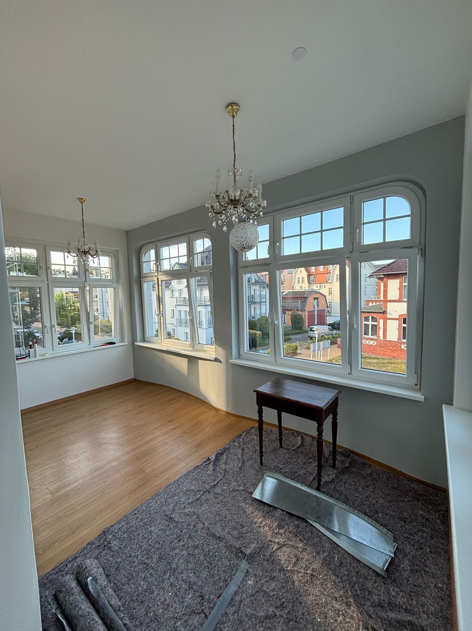 Jasne wnętrze z parkietem i oknami. Podłoga częściowo zabezpieczona matą, widoczny stolik i dekoracyjne żyrandole. Przygotowanie do prac wykończeniowych.