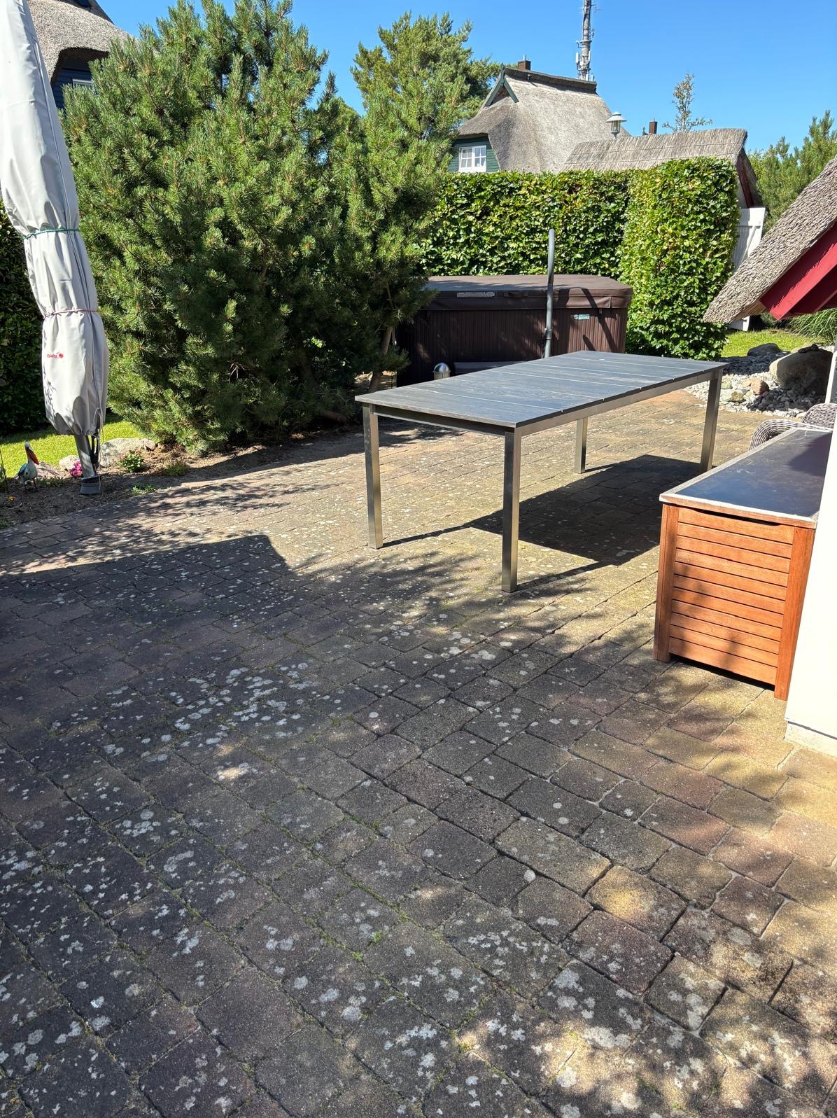 Patio z kostki brukowej z widocznym stołem, krzewami i elementami architektury w tle. Kostka z widocznymi śladami użytkowania i mchu.