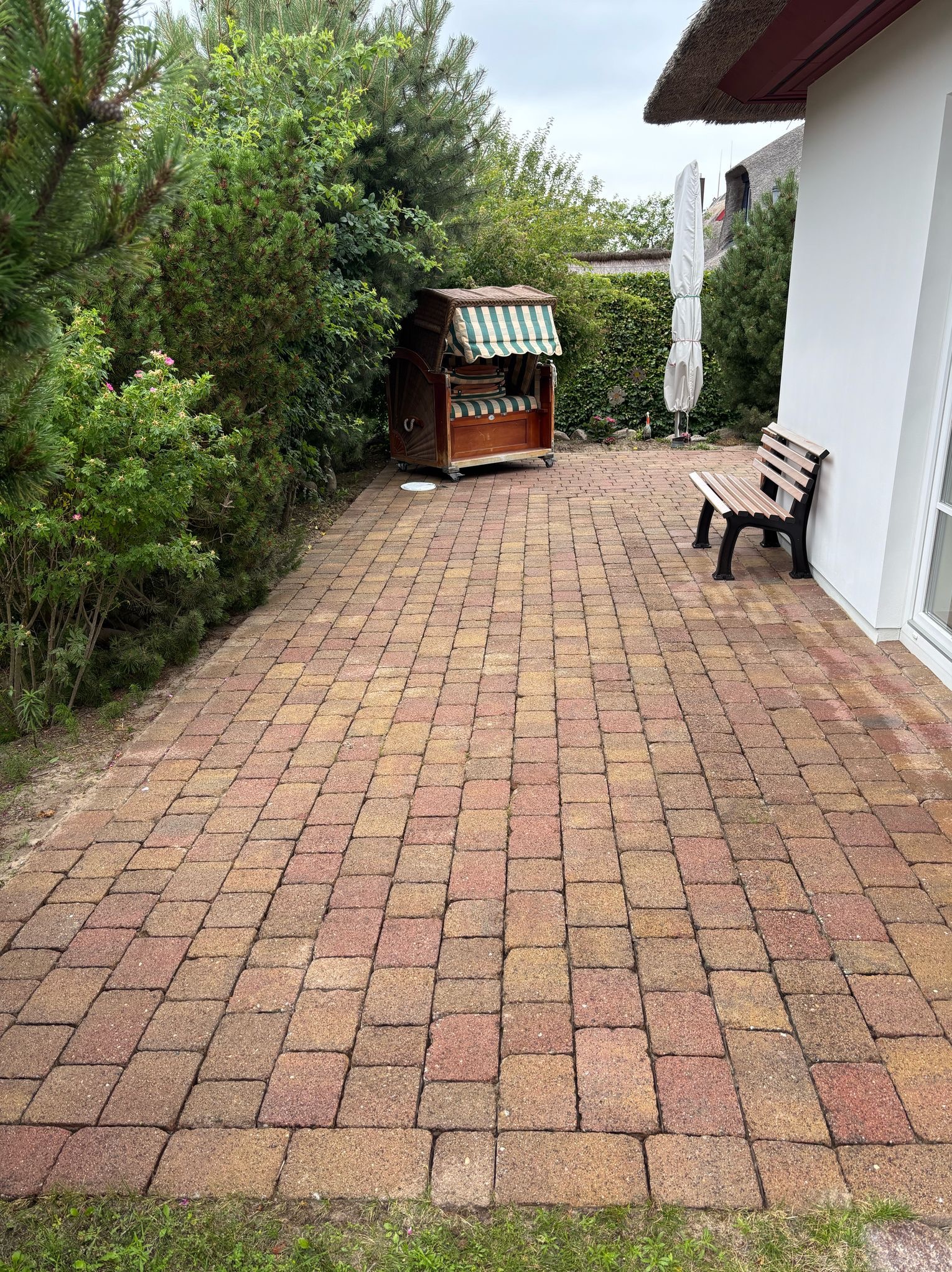 Ułożona kostka brukowa w odcieniach czerwieni i brązu na posesji. W tle widoczna drewniana ławeczka przy białej ścianie domu z strzechą i kosz plażowy.