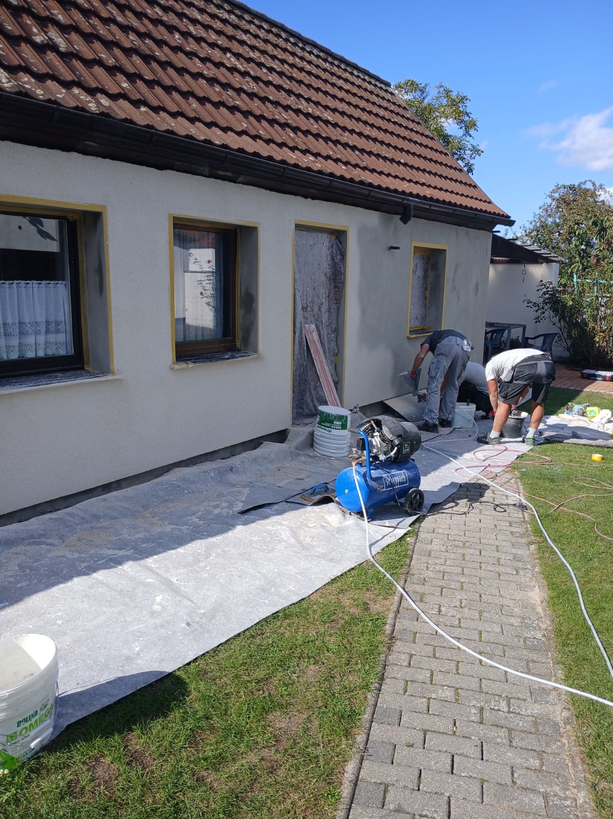 Prace tynkarskie na elewacji domu jednorodzinnego. Dwóch pracowników aplikuje tynk maszynowy. Widoczny kompresor, wiaderka z zaprawą i zabezpieczone okna.
