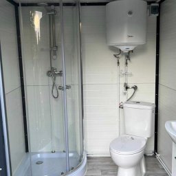 Zuzanna Orzech - Kompaktowa łazienka w kontenerze: kabina prysznicowa narożna, podgrzewacz wody Midea, toaleta i umywalka na podłodze imitującej drewno.