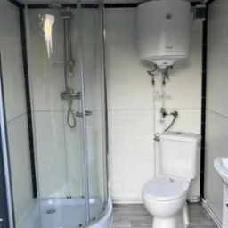 Kompaktowa łazienka w kontenerze: kabina prysznicowa narożna, podgrzewacz wody Midea, toaleta i umywalka na podłodze imitującej drewno.