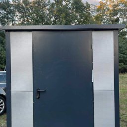 Zuzanna Orzech - Nowoczesna, grafitowa wiata z metalowymi panelami i drzwiami, umieszczona na zewnątrz na trawie. Widoczne elementy montażowe i fragment samochodu.