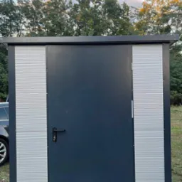 Nowoczesna, grafitowa wiata z metalowymi panelami i drzwiami, umieszczona na zewnątrz na trawie. Widoczne elementy montażowe i fragment samochodu.