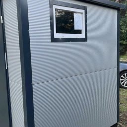 Zuzanna Orzech - Nowoczesna, szara kabina z blachy falistej z oknem i ciemnymi obramowaniami, stojąca na zewnątrz. Widoczny fragment samochodu i drzewa w tle.