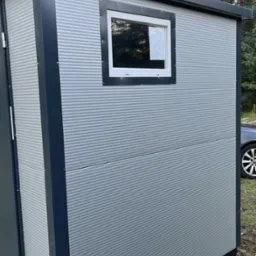 Nowoczesna, szara kabina z blachy falistej z oknem i ciemnymi obramowaniami, stojąca na zewnątrz. Widoczny fragment samochodu i drzewa w tle.