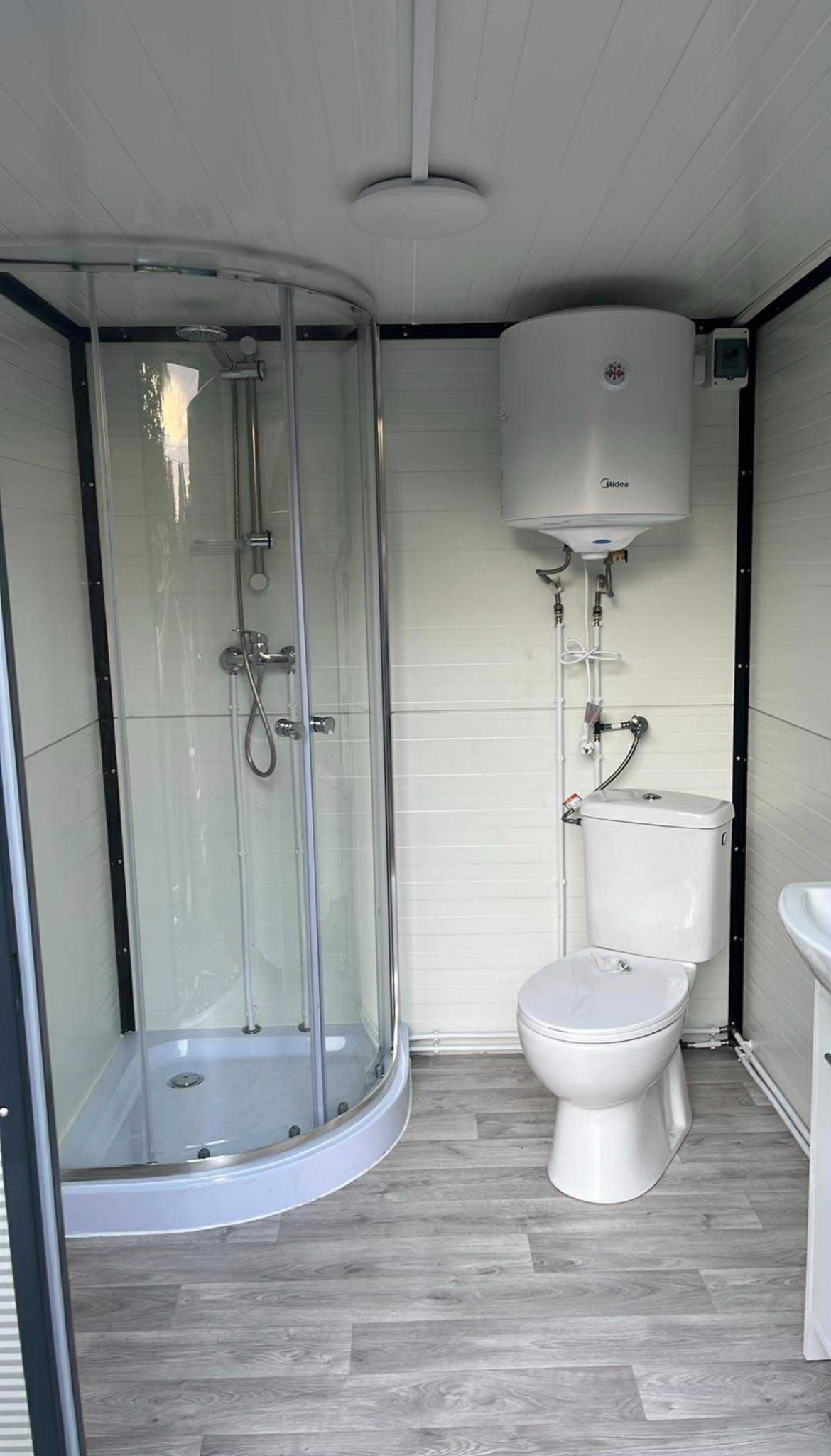 Kompaktowa łazienka w kontenerze: kabina prysznicowa narożna, podgrzewacz wody Midea, toaleta i umywalka na podłodze imitującej drewno.