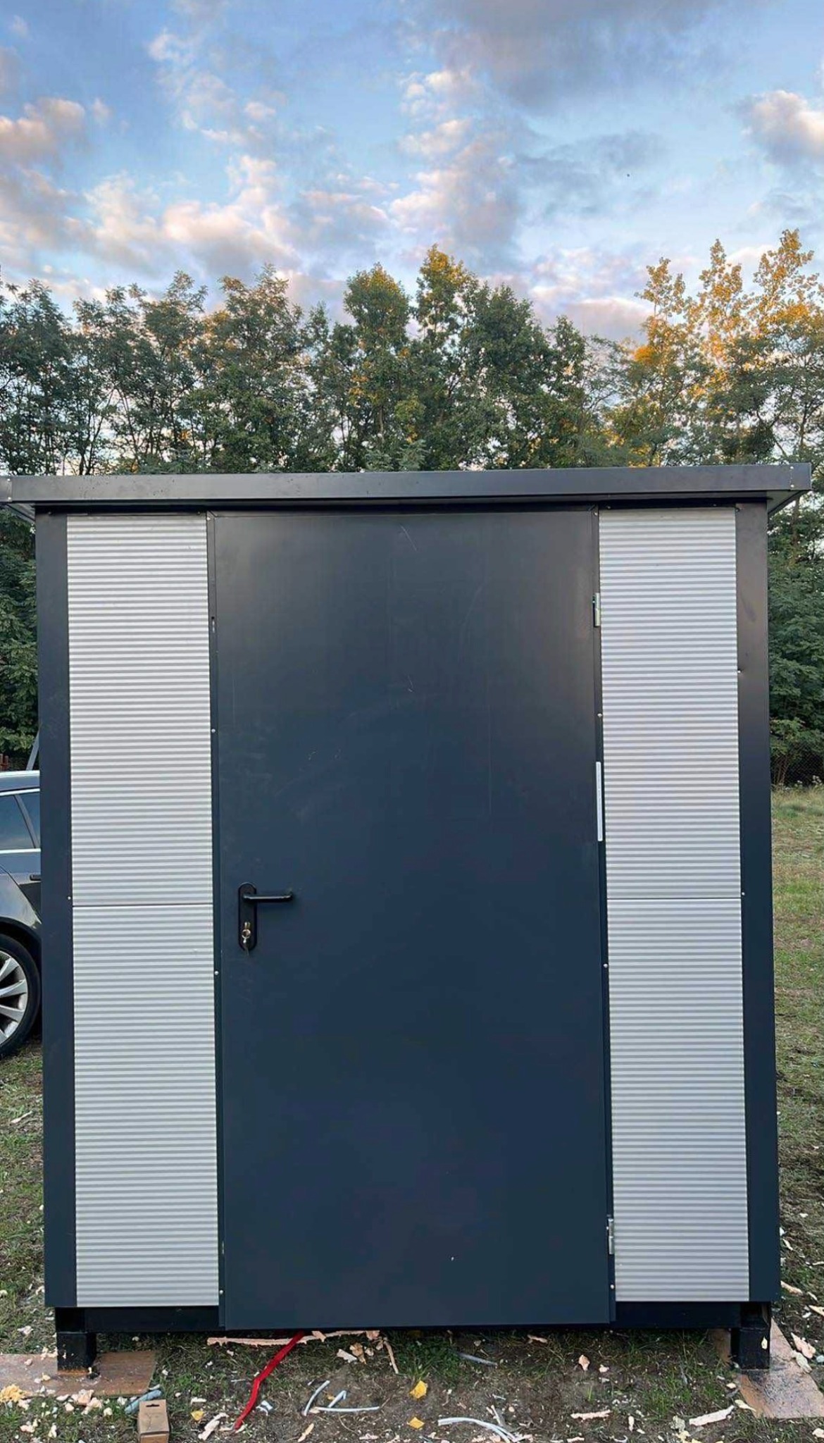 Nowoczesna, grafitowa wiata z metalowymi panelami i drzwiami, umieszczona na zewnątrz na trawie. Widoczne elementy montażowe i fragment samochodu.