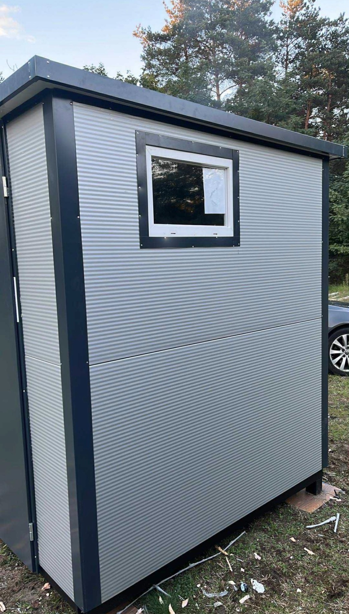 Nowoczesna, szara kabina z blachy falistej z oknem i ciemnymi obramowaniami, stojąca na zewnątrz. Widoczny fragment samochodu i drzewa w tle.