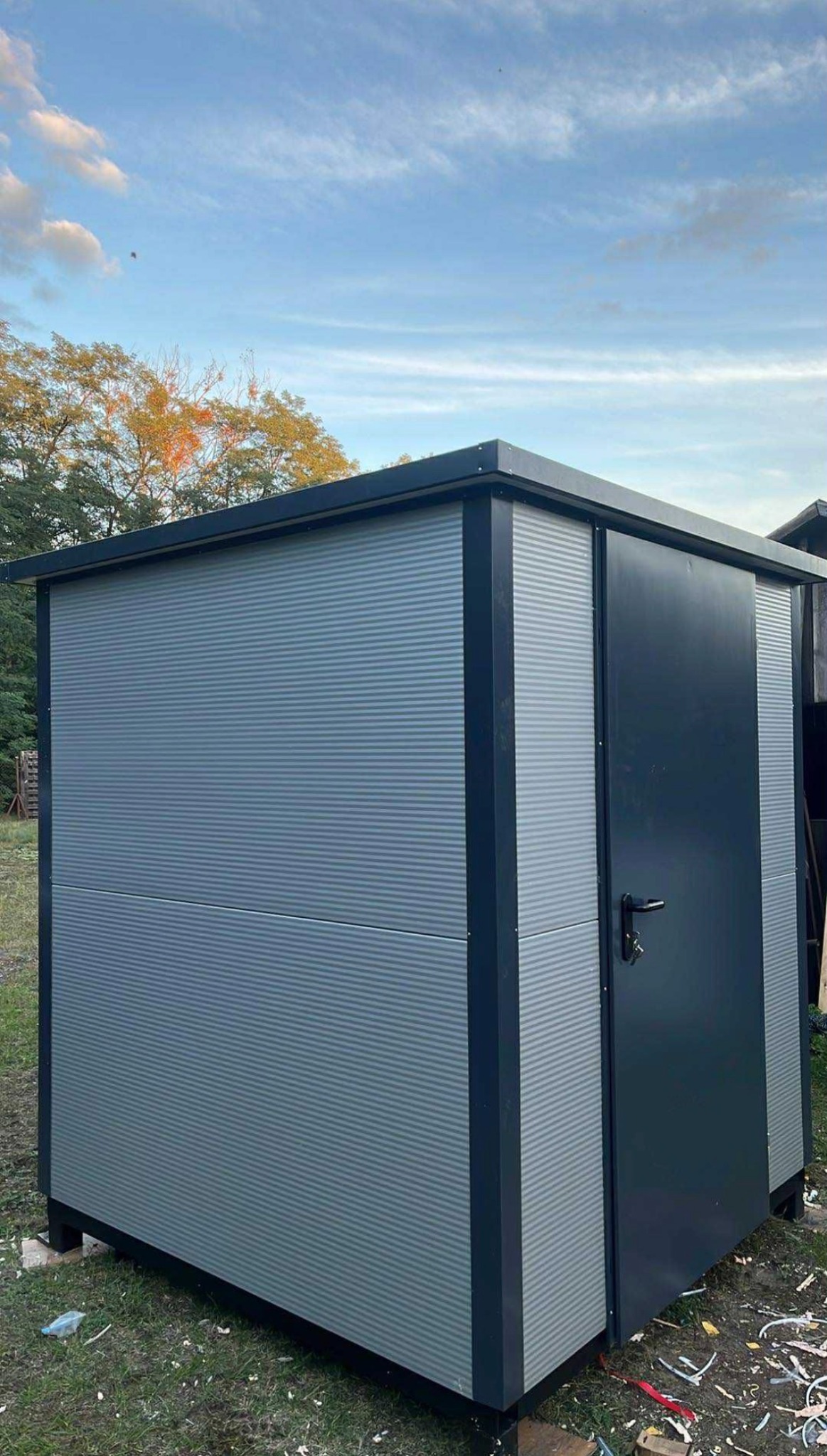 Nowoczesna konstrukcja stalowa w Katowicach: szara, ryflowana obudowa z ciemnymi elementami wykończeniowymi i drzwiami, w otoczeniu zieleni i fragmentów drewna.